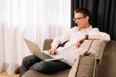 Orta yaşlı gözlüklü bir iş adamı, elinde saatle gri-kahverengi koltukta oturuyor ve dizüstü bilgisayarda çalışıyor. Beyaz gömlekli, koyu renk pantolonlu düşünceli bir erkek ev ofisinden operasyonu büyütüyor..