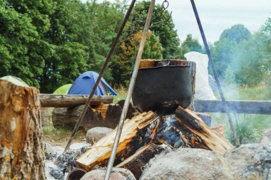 Metal kazanda yemek pişirme işlemi yakacak odunların üzerinde üç ayaklı ayakla asılı, çadırların ve yeşil ormanın karşısında duruyor. Yaz doğasında kamp yapmak için şenlik ateşinde yemek hazırlıyorum..