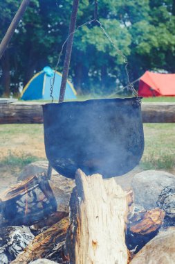 Etrafını çevreleyen metal kazanın buharı içinde üç ayaklı çanak ile farklı renklerde kamp çadırlarına asılmış. Doğada kamp ateşinde yemek pişirmek, mutfağa hazırlanmak. Dikey.