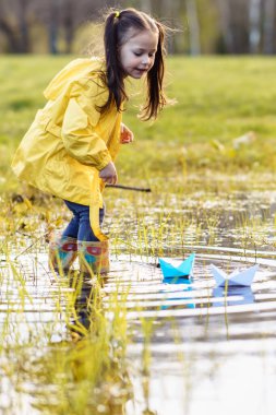 Sarı ceketli, lastik botlu, su birikintisinde duran ve farklı renklerde, suda yüzen iki kâğıt gemiye bakan aktif küçük bir kız. Çocuk hafif tekneleri izliyor, doğada oynuyor. Dikey.