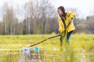 Sarı ceketli, lastik çizmeli, yeşil çimenlikteki su birikintisinde duran ve ağzı açık iki kağıt tekneye bakan hayret verici kadın çocuk. Küçük kız sonbahar günü gemilerle oynuyor..