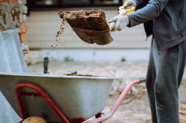 Kır evinin yanında duran, elinde kürek tutan ve toprağı el arabasına atan eldiven giymiş tanınmamış bir insan. Rahat eşofmanlı bahçıvan araziyi araziden temizliyor, toprakla uğraşıyor..