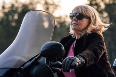 Sonbahar günü deri eldivenli oldukça olgun bir sarışın bisikletini kullanıyor. Hobi motosiklet turizmi. Sağlıklı bir yaşam tarzı. Duygusal rahatlama. Mükemmel bir fiziği var. Turizm ve seyahat.