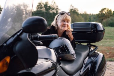 Olumlu duygular. Orta yaşlı mutlu bir şekilde gülümseyen sarışın kameraya bakar, siyah bisikletine yaslanır. Hobi motosiklet turizmi. Sağlıklı bir yaşam tarzı. Mükemmel bir fiziği var. Turizm ve seyahat.