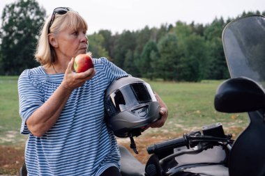 Dişi motorcu. Motosiklet rallisi sırasında motosikletinin yanında elma gibi sarışın, orta yaşlı bir motorcu yiyor. Güçlü ve bağımsız kadınların hobileri. Turizm gezisi. Maceralar. Motosiklet turizmi.