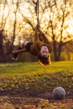 Sonbaharın başında dışarıda oynanan aktif oyunlar. Mutlu genç çocuk doğada, parkta, gün batımının ışının olduğu köyde ev yapımı salıncakta baş aşağı asılı duruyor, futbol yakınlarda yatıyor. Pozitif duygular.