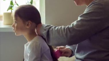 Atış stili zaman ayarlı. Annem küçük kızını evde tarıyor ve ona iki at kuyruğu yapıyor. Anneler ve kızları. Saç bakımı. Annelik. Mutlu bir çocukluk. Sevgi ve ilgi. 4k görüntü.