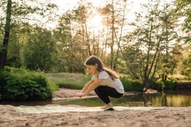 Amaçlı küçük atlet rahat kıyafetler giyip gölün önündeki kumlu sahilde yeşil mindere çömeliyor. Odaklanmış uzun saçlı çocuk gün batımında dışarıda çalışıyor..