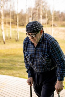 Başörtüsü ve güneş gözlüğü takan yaşlı bir adam dışarıda iki çubuğa yaslanmış yürüyor.