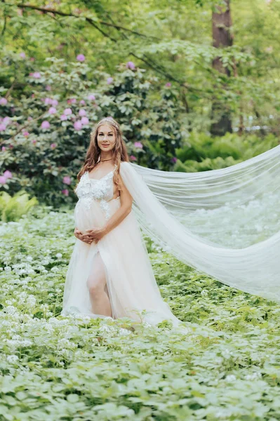 Lüks bir elbise giyen güzel hamile bir kadın çiçek açan bir orman bahçesinde duruyor.