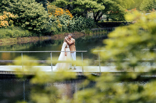 Romantic pregnant couple hugging on wooden bridge in green blooming nature