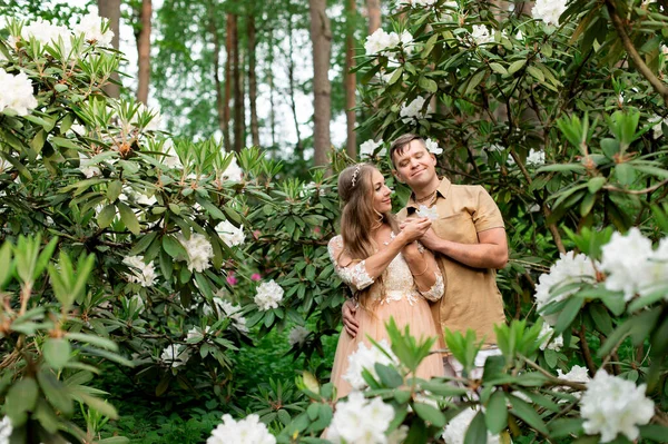ロマンチックな妊娠中のカップルの花の庭で抱擁 ホワイトシャクナゲ公園の将来の両親 — ストック写真