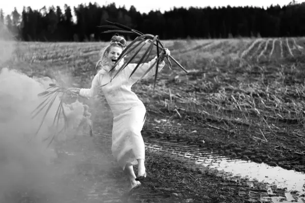 Happy woman in white dress with smoke bomb and reeds dancing on muddy field