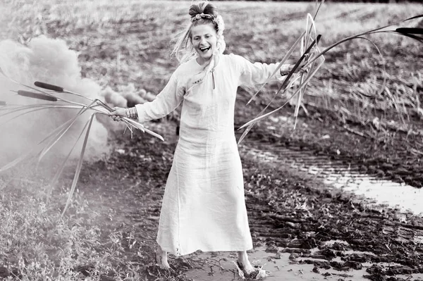 Happy woman in white dress with smoke bomb and reeds dancing on muddy field