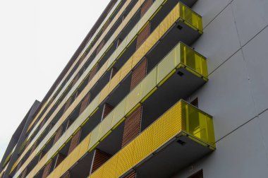 Gri ve sarı tonlarında modern bir apartmanın dış cephesi, metal balkon korkulukları ve duvar panellerinde kahverengi tahta panjurlar.