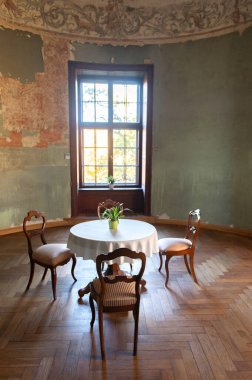 Yuvarlak masa ve eski bir ortaçağ şatosunun odalarında üç sandalye. Pencereden gelen doğal gün ışığı.