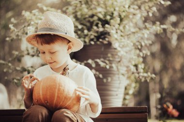 Tahta merdivenlerde oturan, elinde bir balkabağı tutan, hasır şapka takan, arka planda sonbahar bitkileri olan genç bir çocuk.