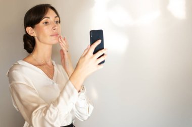 Beyaz bluzlu zarif bir kadın açık gri arka planda akıllı telefonuyla selfie çekiyor..