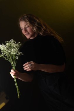 Uzun kıvırcık saçlı, gülümseyen ve arka planda beyaz çiçekleri kokan, Sevgililer Günü 'nü veya yıldönümünü kutlayan genç bir kadının stüdyo fotoğrafı. Reklam kavramı.