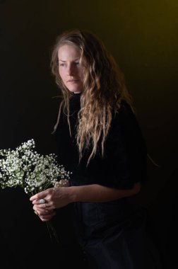 Uzun kıvırcık saçlı, gülümseyen ve arka planda beyaz çiçekleri kokan, Sevgililer Günü 'nü veya yıldönümünü kutlayan genç bir kadının stüdyo fotoğrafı. Reklam kavramı.