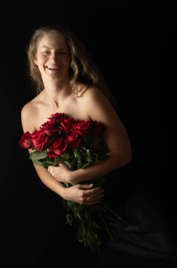 Stüdyonun arka planında çıplak omuzları ve kırmızı gülleri olan seksi bir kadın. Güzel stüdyo portresi. Siyah üzerine izole edilmiş kırmızı güllü şehvetli bir model. Makyajsız, olgun, doğal bir kadın..