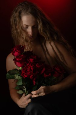Stüdyonun arka planında çıplak omuzları ve kırmızı gülleri olan seksi bir kadın. Güzel stüdyo portresi. Siyah üzerine izole edilmiş kırmızı güllü şehvetli bir model. Makyajsız, olgun, doğal bir kadın..