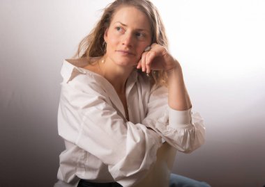 Beyaz gömlekli güzel bir kadın stüdyo koltuğunda poz veriyor. Beyaz stüdyo arka planında uzun kıvırcık saçlı mutlu bir yetişkin kadın. Doğal duyguları olan etkileyici bir portre..