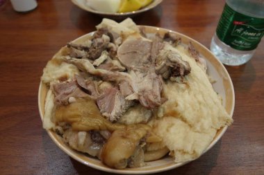 Tavuk ve muhtemelen domuz eti de dahil olmak üzere çeşitli et kesimlerinin yer aldığı geleneksel bir tabak et, yumuşak ekmek veya hamurdan oluşan bir yatakta servis ediliyor. Tabak sağlıklı görünüyor ve arka planda bir şişe suyla ahşap bir masada sunuluyor..