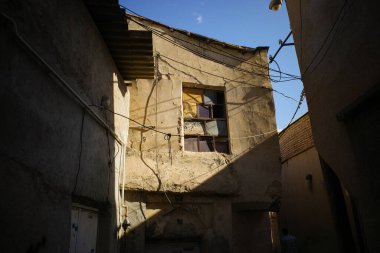 Eski, yıpranmış alçılı binalar arasında dar bir ara sokak. Kırık camlı bir pencere görülebilir ve elektrik kabloları çaprazlama kesişir. Sahne doğal ışıkla aydınlatılır, yere gölgeler düşer..