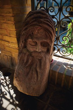 A carved wooden sculpture depicting a bearded face with deep-set eyes and a textured beard, set against a brick wall with wrought iron details. clipart