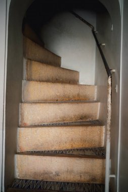 Yukarı doğru çıkan aşınmış bir merdiven, dokulu basamaklar ve basit bir korkuluk. Duvarlar sade ve ışık yumuşak, sakin bir atmosfer yaratıyor..