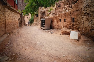 Yıpranmış taş duvarlarla çevrili kırsal bir köyden geçen dar bir toprak yol. Arka planda toprak tonları ve yeşilliklerle çevrili eski bir metal dolap yerde duruyor..