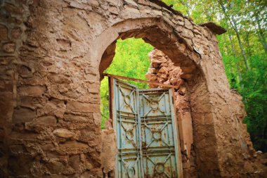 Paslı mavi kapısı kısmen açık, yemyeşil yapraklarla çevrili eski, yıpranmış bir taş kemer yolu. Harabeler tarihi bir yeri işaret ediyor, doğa yapıyı geri istiyor..