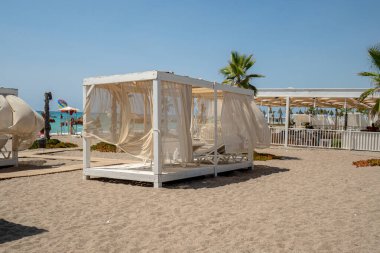 Tipik tropikal atmosfer, plaj tatili, güneşli bir günde palmiye ağaçları, baldaquin, şezlonglar ve arkada okyanus olan bir tatil köyünde dinlenmek.