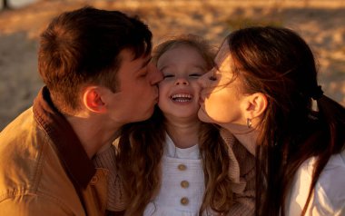 Genç anne ve baba, hafta sonu gün batımında kumsalda tatlı kızlarını öpüyor ve eğleniyorlar..