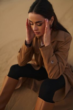 Kumda oturan yağmurluk ve çizme giymiş güzel bir kadın. Sonbaharda kumlu bir sahilde zarif bir kadın..