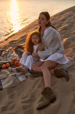 Gün batımında kumlu bir sahilde piknikte annesiyle birlikte battaniye örüp eğlenen ve hafta sonu dinlenen küçük tatlı bir kız..