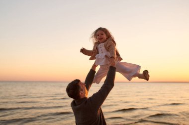 Tatlı kıvırcık bir kızı olan genç bir baba gün batımında deniz kenarında kumsalda yürüyor ve eğleniyor..