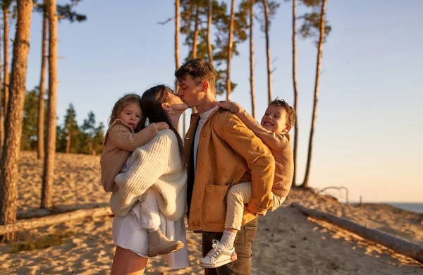 A young family with two children on their backs kissing on a sandy beach against the backdrop of pine trees at sunset over the weekend relaxing and having fun.