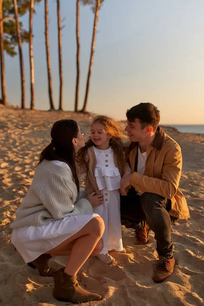 Young mom and dad kiss their cute daughter on a sandy beach against the backdrop of pine trees at sunset on the weekend relaxing and having fun.