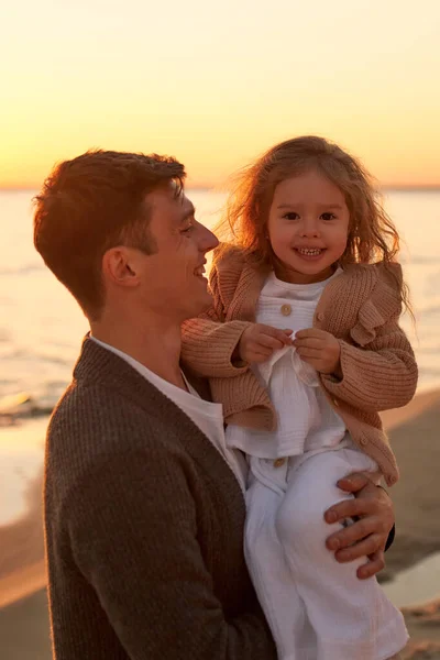 A young dad with a cute curly daughter walking on a sandy beach near the sea at sunset having fun and playing.