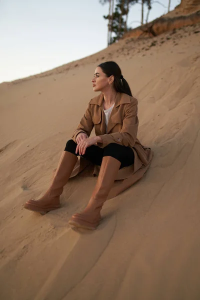 美しい女性のレインコートやブーツの砂の上に座っている 秋の砂浜で優雅な女性 — ストック写真