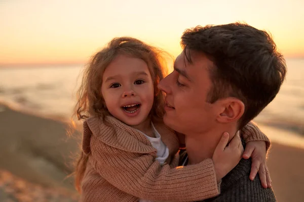 A young dad with a cute curly daughter walking on a sandy beach near the sea at sunset having fun and playing.