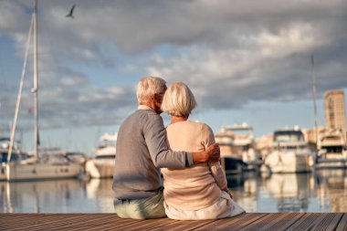 Yaşlı, zeki, gri saçlı, aşık bir çift lüks yatları ve tekneleri olan bir rıhtımda oturmuş deniz manzarasını izlerken romantik vakit geçiriyorlar..