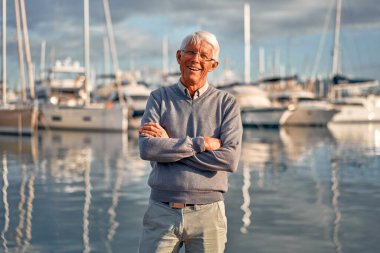 Yaşlı, zeki, gri saçlı bir adam rıhtımda yürüyor lüks yatları ve tekneleri var, deniz manzarasını seyrediyor ve dinleniyor..
