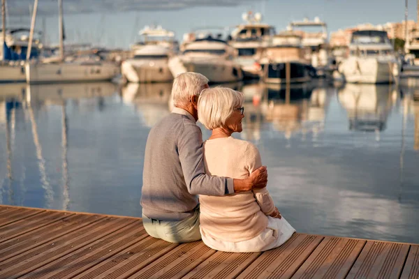 stock image An elderly, intelligent, gray-haired couple in love are sitting on a pier in a seaport, having a romantic time admiring the seascape.