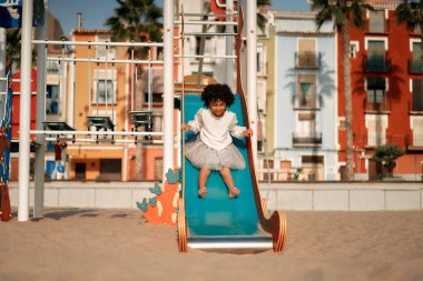 Beyaz elbiseli, afro kıvırcık saçlı tatlı Afro-Amerikalı kız oyun parkında oynuyor, kumlu sahilde kaydırağın üzerinde sahil evlerinin arka planına doğru gidiyor..