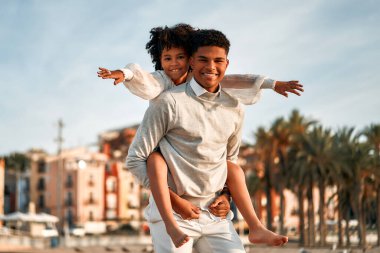 Afrika kökenli Amerikalı bir aile deniz kenarındaki kumlu sahilde eğleniyor. Sırtında küçük şirin kız kardeşiyle sahilde sahil evlerinin arka planında oynayan bir ağabey..