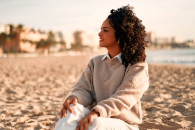 Güzel Afro kıvırcık saçlı, sıcacık kazaklı Afro-Amerikalı kadın deniz kenarında kumlu bir sahilde oturuyor ve deniz manzarasına bakıyor..