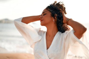 Beyaz bluzlu afro bukleli güzel Afrikalı Amerikalı kadın deniz kenarında kumlu bir sahilde güneşin tadını çıkarıyor..